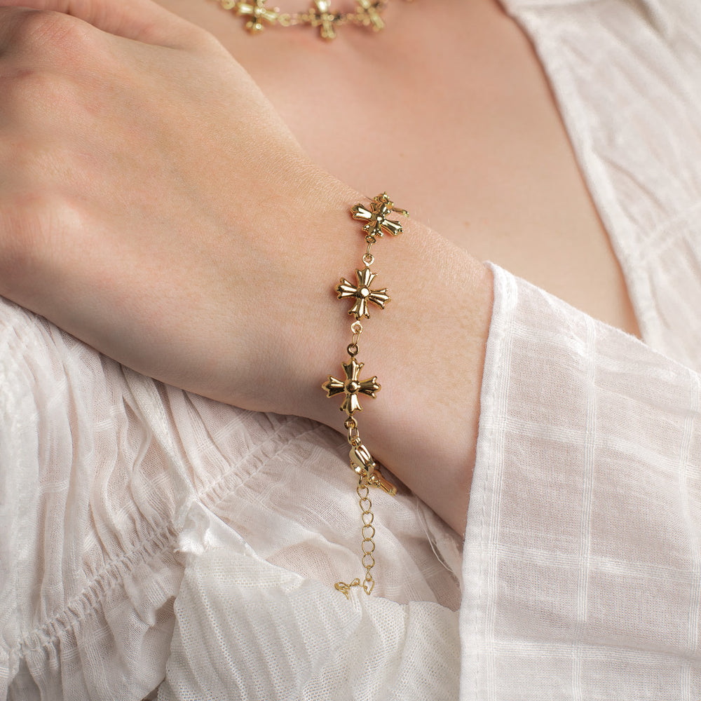 
                  
                    Gold dainty bracelet with cross designs on a person wearing a white blouse.
                  
                