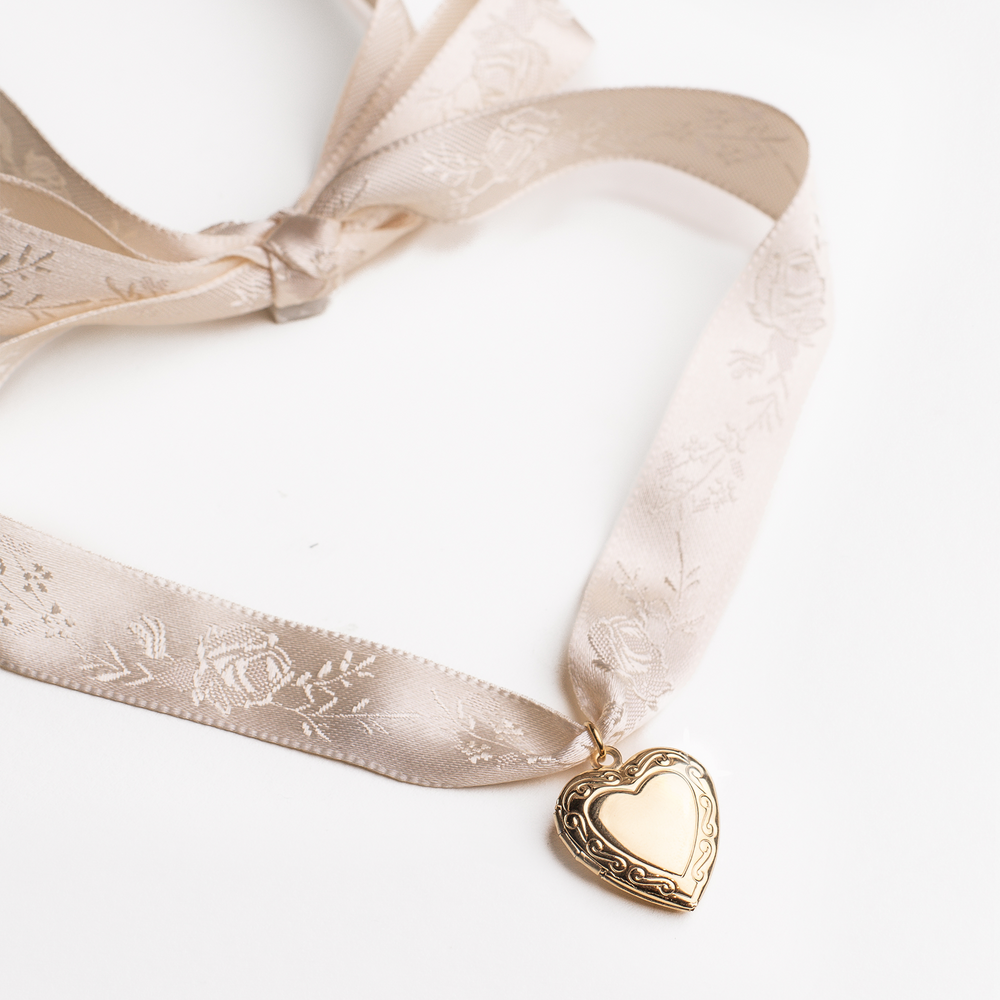 
                  
                    Gold heart shaped locket pendant on a beige ribbon with floral details against a white background
                  
                
