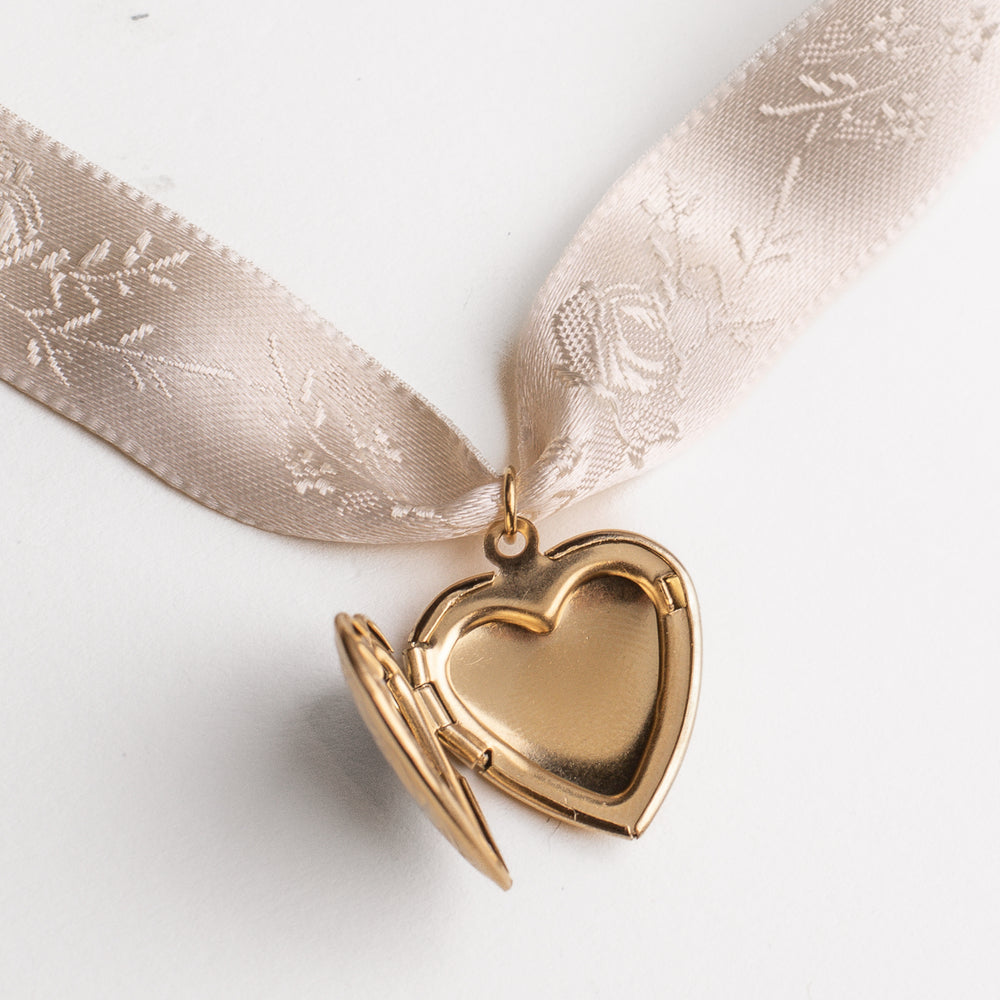 
                  
                    Vintage heart locket gold pendant on a cream satin ribbon against a white background
                  
                